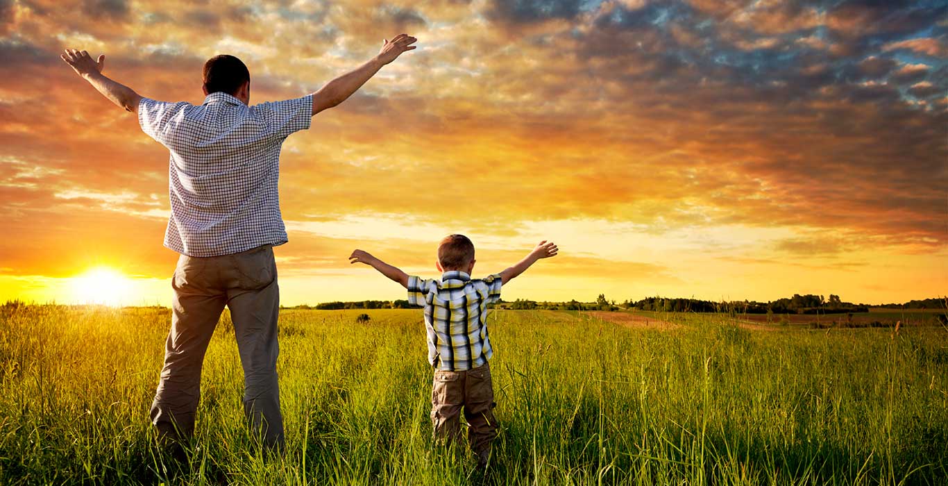 A father and child raising their hands in an open field overlooking the green vegetation.